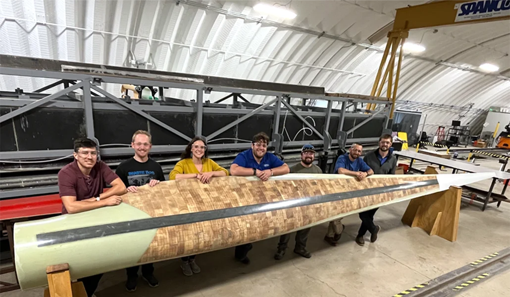 seven people pose beind a large wind turbine blade in a manufacturing facility